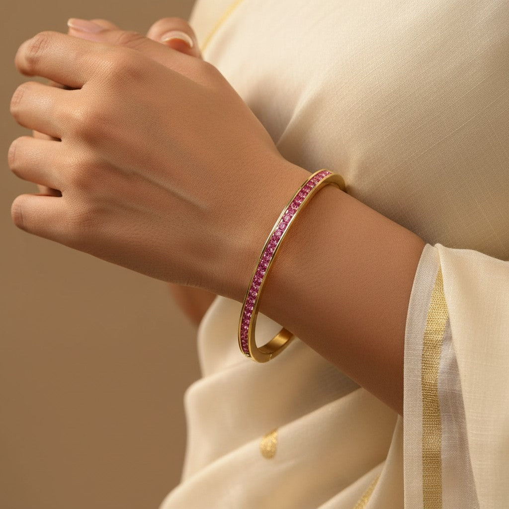Aurevia Fashion Jewellery gold-toned bangle featuring vibrant pink stones worn on a woman's wrist with a white saree.