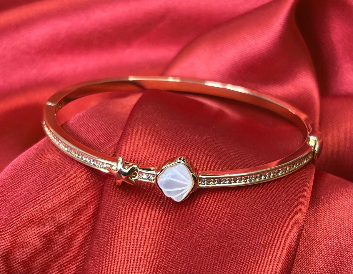 Aurevia Fashion Jewellery gold-tone bangle featuring a mother of pearl shell inlay and starfish accent on red satin.