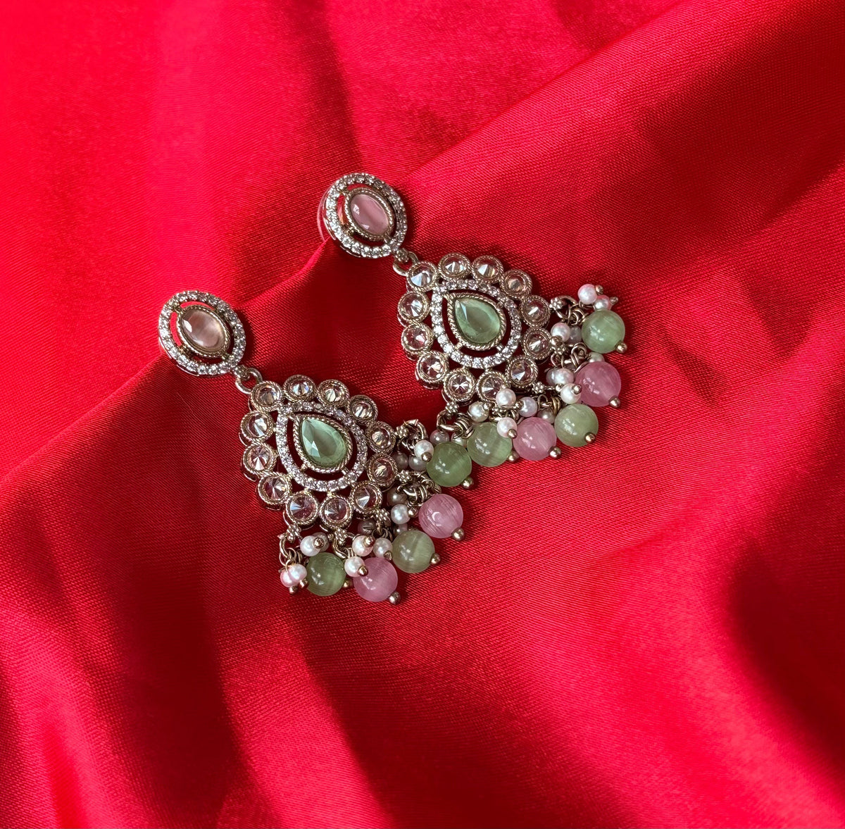 Aurevia Fashion Jewellery Noor multi-color kundan drop earrings with pastel gemstones on a vibrant red silk background.